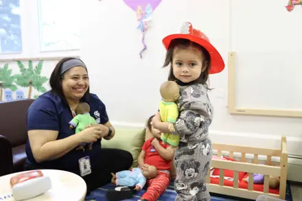 toddler girl playing dress up with teacher and holding a doll